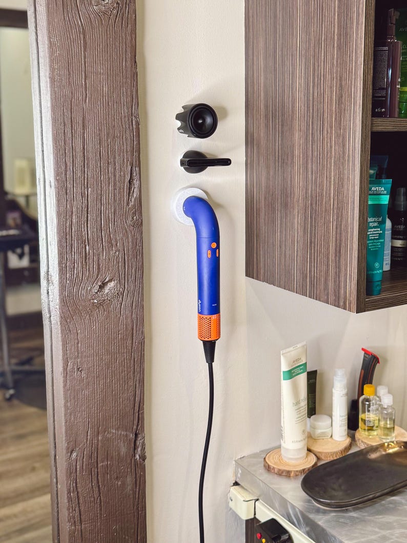 May include: A blue and orange hair dryer with a long cord is mounted on a white wall. The hair dryer is attached to a white wall mount. The hair dryer is in a salon setting.