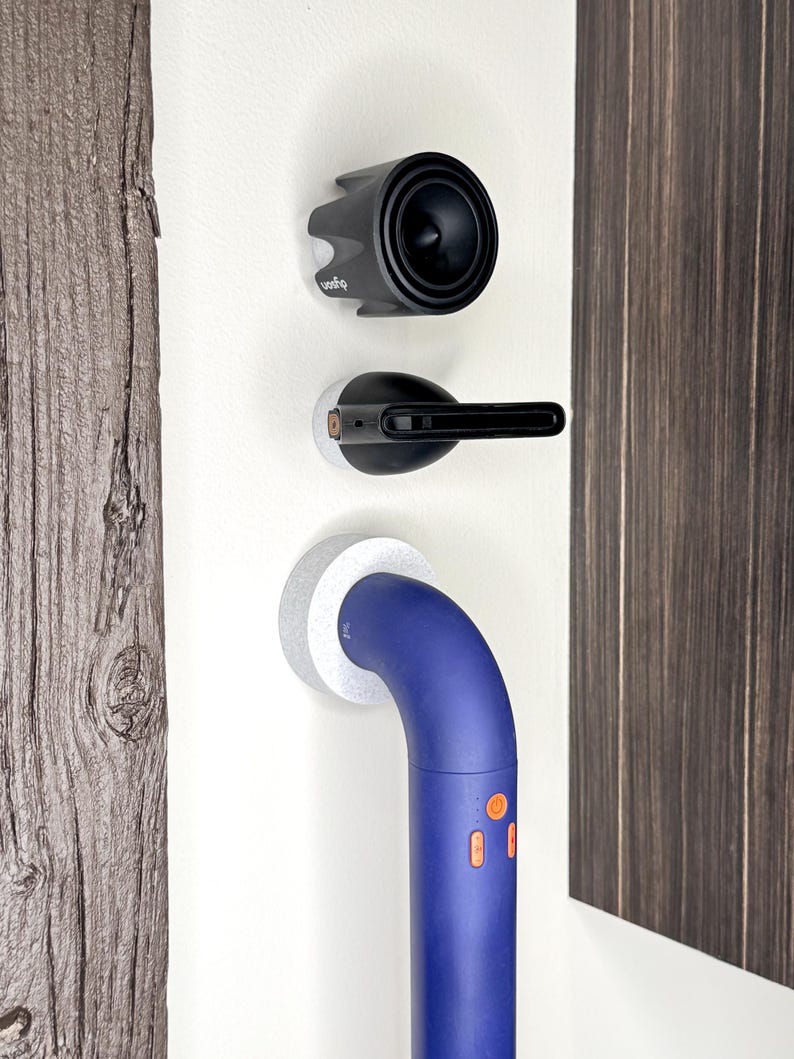 May include: A white wall with three objects mounted on it. The top object is a black speaker with a white logo that says "Q-Sound". The middle object is a black and silver device with a white base. The bottom object is a blue and white handrail with two orange buttons.