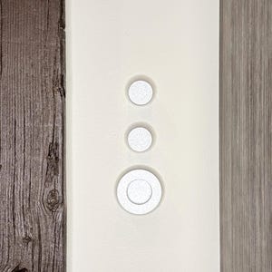 May include: Three white round light switch covers are mounted vertically on a white wall. The switch covers are slightly raised from the wall.