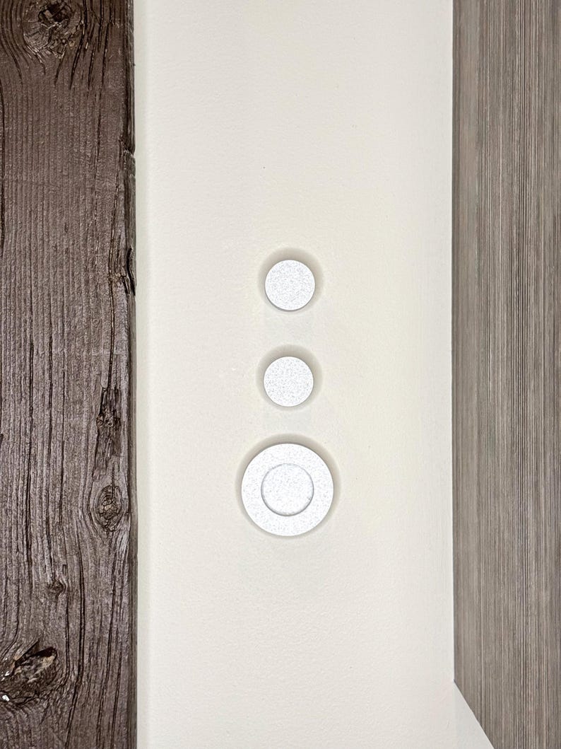 May include: Three white round light switch covers are mounted vertically on a white wall. The switch covers are slightly raised from the wall.