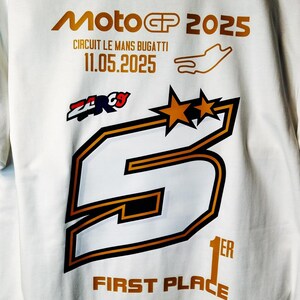 Peut inclure: T-shirt blanc avec un motif doré et noir. Le t-shirt affiche le texte "MotoGP 2025" et "Circuit Le Mans Bugatti 11.05.2025". Un grand chiffre "5" est affiché avec deux étoiles et les mots "First Place".
