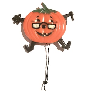 May include: A vintage orange pumpkin-shaped Halloween decoration with black arms and legs, wearing glasses and a smiling face. The pumpkin has a green stem and a string attached, perfect for hanging.