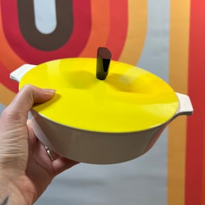 May include: A vintage, cream-colored casserole dish with a bright yellow lid. The lid features a dark brown, geometric handle. The dish has white handles on either side. The background has a retro, colorful design.
