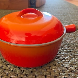 May include: Orange enameled cast iron saucepan with a wooden handle and a lid. The saucepan has a gradient of orange and red.
