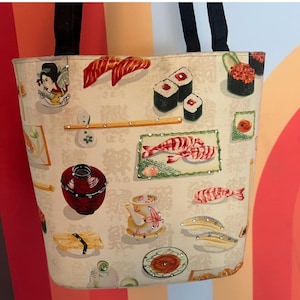 May include: Beige tote bag featuring a sushi-themed pattern with illustrations of various sushi rolls, sashimi, and Japanese teacups. The bag has black handles and is embellished with small, decorative rhinestones.
