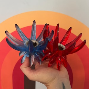 May include: Two decorative candle holders in the shape of starbursts. One is transparent blue, the other is transparent red. Each has a silver-colored metal cup in the center for holding a candle. The background features a retro rainbow design.