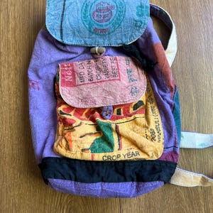 May include: A handmade backpack with a purple body, a blue flap, and a colorful front pocket. The backpack features text and a wooden toggle closure. The straps are beige and the pocket has a yellow and orange design.