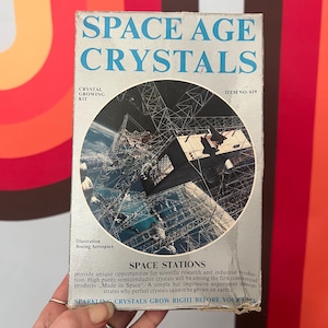 May include: A vintage "Space Age Crystals" crystal growing kit box. The box is light blue with the words "SPACE AGE CRYSTALS" in large blue letters. The box features an illustration of a space station and the text "CRYSTAL GROWING KIT".