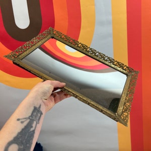 May include: A gold-toned rectangular tray with an ornate, decorative frame and a mirrored surface.