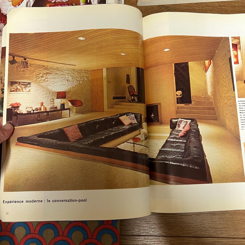 May include: A large, black leather sectional sofa with a wooden frame sits in a modern living room with a wood-paneled ceiling. The sofa is arranged in a U-shape, creating a conversation pit. The room is decorated with a stone wall and a large window. The text "Experience moderne: le conversation-pool" is printed below the image.