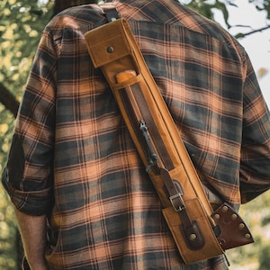 May include: A tan canvas and brown leather shoulder sling bag featuring a long vertical zippered compartment, a buckled strap closure, and a side pocket containing a small tool with a wooden handle, worn diagonally across the back.