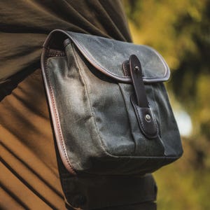 Może przedstawiać: Ciemnozielona torba z woskowanego płótna z klapką i brązowymi skórzanymi wykończeniami. Torba jest zabezpieczona skórzanym paskiem i metalowym guzikiem. Torba jest przymocowana do ubrania osoby.