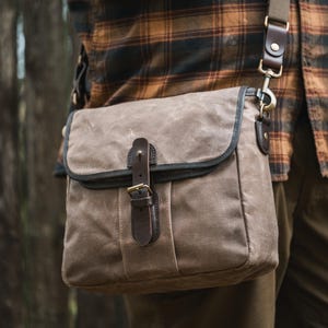 Könnte beinhalten: Eine Messenger-Tasche aus gewachstem Canvas in einem gedämpften Braunton, mit Lederriemen und Schnallenverschluss. Die Tasche hat eine dunkelgrüne Einfassung und einen braunen Leder-Schulterriemen mit Messingbeschlägen.