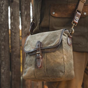 May include: Olive green waxed canvas messenger bag with brown leather accents. The bag features a flap closure with a buckle, a shoulder strap, and a brown leather trim. The bag is worn over a matching olive green jacket.