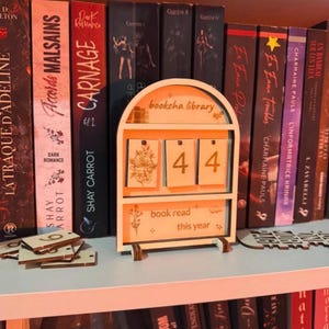 Puede incluir: Un rastreador de libros de madera con el texto "bookcha library" y "book read this year". El rastreador muestra el número "44". El rastreador está en un estante con una colección de libros.