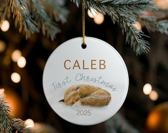 May include: A white, circular Christmas bauble with the name "CALEB" in gold. The words "First Christmas" are written in a script font, and a sleeping fox illustration is featured. The year "2025" is printed below the fox.