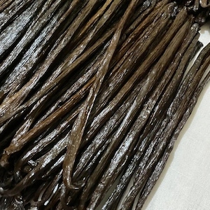 May include: A close-up image of a bundle of dark brown vanilla beans. The beans are arranged in a neat pile, with their tips pointing in different directions. The beans are shiny and smooth, with a slightly oily texture.