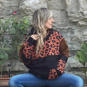Può includere: Una felpa con cappuccio marrone e nera con stampa leopardata con cappuccio e maniche nere. La felpa con cappuccio è abbinata a jeans blu e scarpe da ginnastica bianche. La persona è seduta su una panchina di legno davanti a un muro di pietra.