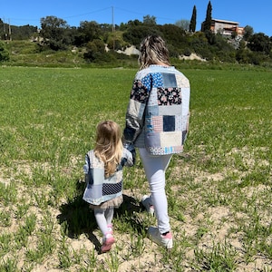 May include: A woman and child wearing patchwork jackets walk through a field of green grass under a clear blue sky. The jackets feature a variety of fabric patterns, including floral and solid squares. The child wears a skirt and pink shoes.