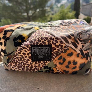 Puede incluir: Un neceser rectangular de patchwork con asa marrón. La bolsa presenta estampados de animales y florales en tonos marrón, naranja, verde y negro. Una pequeña etiqueta negra con texto blanco está cosida en la parte delantera.
