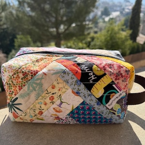 Puede incluir: Un neceser acolchado y rectangular con cierre de cremallera y asa marrón. La bolsa presenta un diseño de patchwork con varios estampados florales y geométricos en tonos rojos, azules, amarillos y verdes. Los textos "Hem" y "pinkstudio" son visibles.