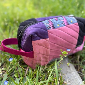 Può includere: Un beauty case rettangolare trapuntato con cerniera nera e manico magenta. La borsa presenta pannelli di tessuto nero, viola, rosa e blu con una stampa animalier rosa ripetuta. La borsa è nell'erba verde.