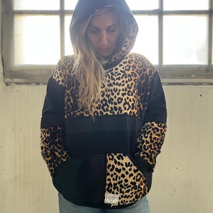 Peut inclure: Sweat à capuche noir avec un imprimé léopard sur le devant et les manches. Le sweat-shirt a une poche kangourou et une capuche noire. Le modèle porte un short en jean.