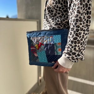 Op de afbeelding: Een denim patchwork clutch met een verscheidenheid aan kleurrijke stoffen vierkanten. De clutch heeft een gewatteerd ontwerp en een donkerblauwe denim rand. De achtergrond toont een persoon die een hoodie met luipaardprint en een beige broek draagt.