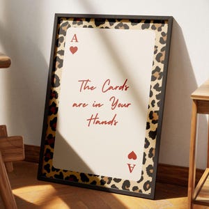 May include: Framed art print featuring a playing card design. The central image is a cream-colored Ace of Hearts card with the text "The Cards are in Your Hands" in red script. The frame has a leopard print border.