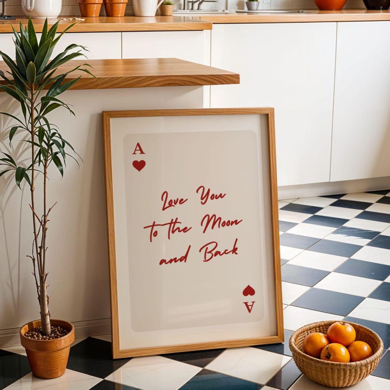 May include: A framed print featuring the Ace of Hearts card design with the text "Love You to the Moon and Back" in red script. The print is in a wooden frame and leans against a white wall. A small potted plant and a basket of oranges are in the foreground.