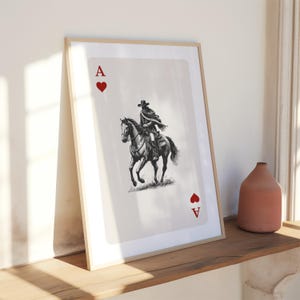 K&ouml;nnte beinhalten: Gerahmter Kunstdruck der Herz-Ass-Spielkarte mit einem Cowboy auf einem Pferd. Die Schwarz-Wei&szlig;-Illustration ist vor einem cremefarbenen Hintergrund platziert, mit roten Herzsymbolen in den Ecken. Der Rahmen hat eine helle Holzfarbe.