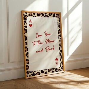 May include: A framed print featuring a playing card design with a leopard print border. The card is an Ace of Hearts, with the text "Love You to the Moon and Back" in red script. The frame is a light brown color.