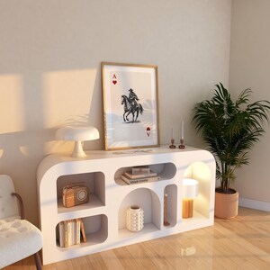 K&ouml;nnte beinhalten: Ein wei&szlig;er Schrank im modernen Stil mit offenen Regalen und geschwungenem Design. Der Schrank zeigt einen gerahmten Druck einer Spielkarte, eine Lampe, B&uuml;cher und dekorative Gegenst&auml;nde. Eine Topfpflanze und ein Stuhl sind ebenfalls sichtbar.