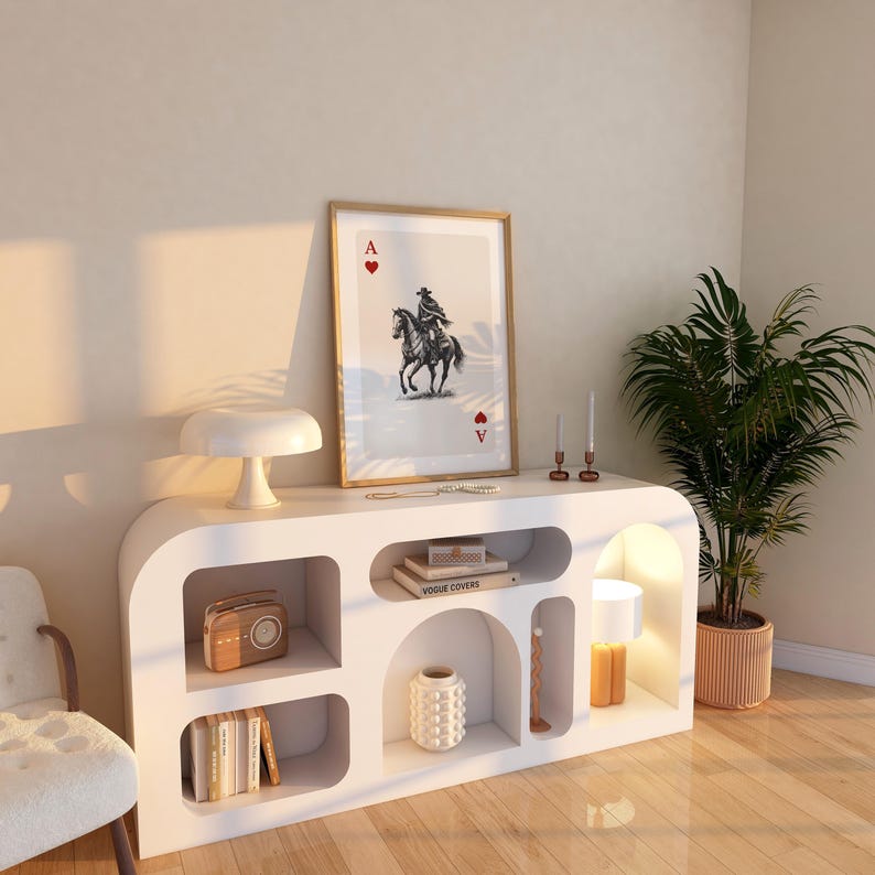 K&ouml;nnte beinhalten: Ein wei&szlig;er Schrank im modernen Stil mit offenen Regalen und geschwungenem Design. Der Schrank zeigt einen gerahmten Druck einer Spielkarte, eine Lampe, B&uuml;cher und dekorative Gegenst&auml;nde. Eine Topfpflanze und ein Stuhl sind ebenfalls sichtbar.