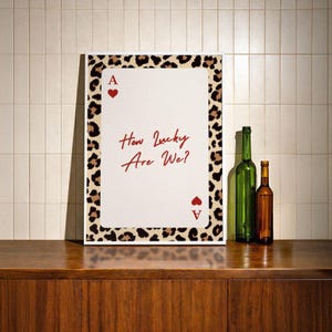 May include: A framed print featuring a playing card design with the text "How Lucky Are We?" in red script. The card has a leopard print border and the Ace of Hearts symbol. Two bottles, one green and one brown, are on a wooden surface.