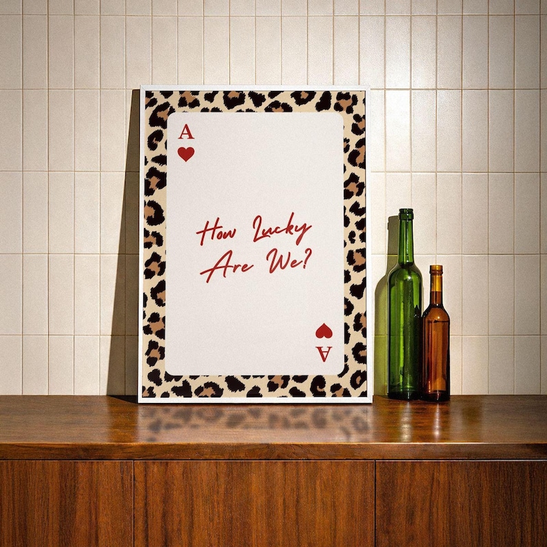 May include: A framed print featuring a playing card design with the text "How Lucky Are We?" in red script. The card has a leopard print border and the Ace of Hearts symbol. Two bottles, one green and one brown, are on a wooden surface.