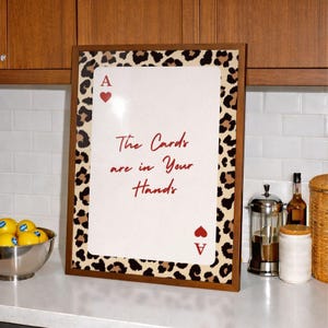 May include: A framed print with a leopard print border and the text "The Cards are in Your Hands" in red script. The print features the Ace of Hearts card symbol. The frame is brown.