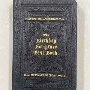 Puede incluir: Un libro encuadernado en cuero negro con letras doradas. El título es "The Birthday Scripture Text Book." La portada presenta adornos decorativos y el texto "Pray one for another. Jas. V. 16" y "Hear my prayer, O Lord. Ps. XXXIX. 12."