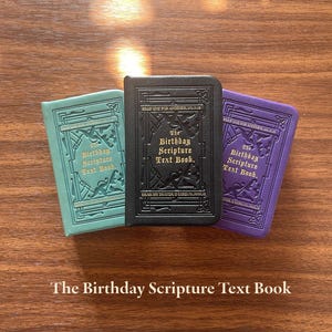以下が含まれることがあります： 「The Birthday Scripture Text Book」というタイトルの小さな本が3冊。色はティール、黒、紫です。各表紙にはエンボス加工の装飾的な縁取りと金色の文字が施されています。黒い本が他の2冊の間に置かれ、木製の表面に並んでいます。