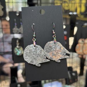 May include: A pair of silver-toned, flat, circular earrings shaped like the Millennium Falcon spaceship. The earrings feature intricate, etched mechanical detailing on the surface and are attached to iridescent, rainbow-colored metal hooks.