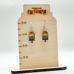 May include: A pair of rainbow popsicle earrings with silver hooks. The earrings are displayed on a wooden stand with the text "FORGED BY FANDOMS". The stand has measurements in inches.