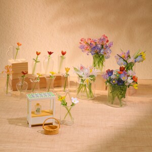 Peut inclure: Une collection de petits vases en verre remplis de fleurs artificielles colorées. Les vases sont disposés sur une surface en bois avec une petite table blanche et un panier.