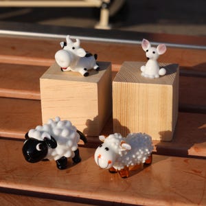 May include: Four glass animal figurines on wooden blocks. A white cow with black spots, a white sheep with black eyes, a white sheep with brown eyes, and a white rabbit with pink ears.