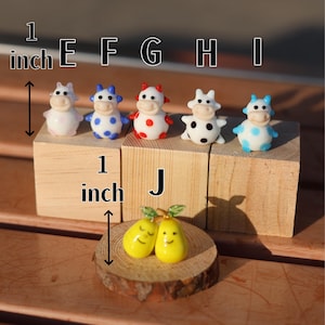 May include: Six miniature glass cow figurines with different colored polka dots, each measuring 1 inch tall. The cows are arranged on wooden blocks labeled E through I. A pair of yellow glass pears are on a wooden slice labeled J.