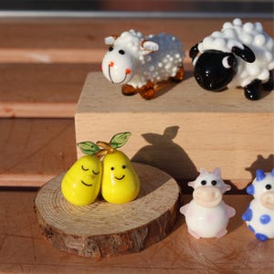 May include: A set of glass figurines, including two yellow pears with smiling faces, a white cow with black eyes, a blue cow with white spots, and two white sheep with black eyes. One sheep has a black face.