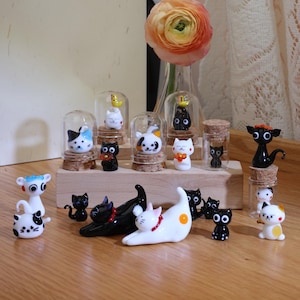 Op de afbeelding: Een verzameling glazen kattenbeeldjes in verschillende poses en kleuren, waaronder zwart, wit en oranje. De beeldjes worden tentoongesteld op een houten plank met een bloemige achtergrond.