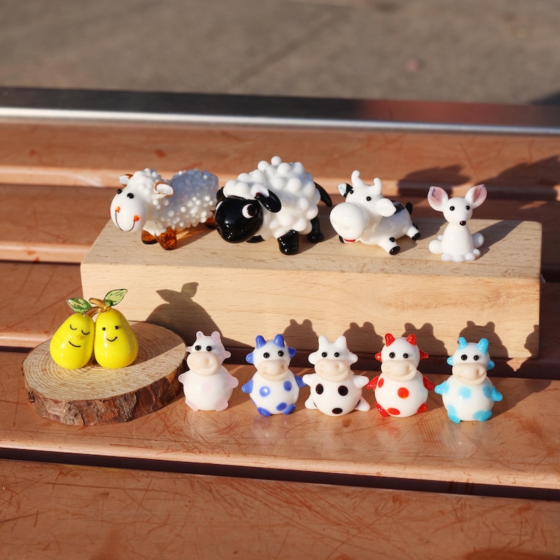 May include: A collection of glass animal figurines, including cows, sheep, and a rabbit. The figurines are painted in various colors, including black, white, blue, red, and yellow. The figurines are arranged on a wooden surface.