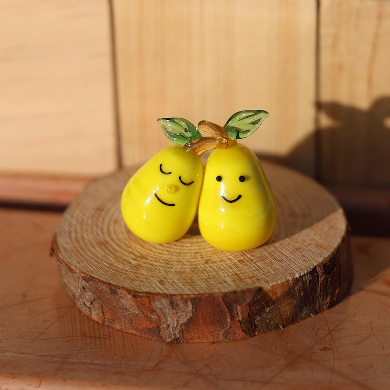 May include: Two yellow glass pears with painted faces, one with closed eyes and a smile, the other with open eyes and a smile, on a wooden base.