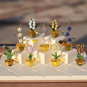 May include: A collection of miniature glass flower arrangements in various colors, including white, yellow, pink, blue, and green. The flowers are displayed on a tiered white stand with a wooden base. Each arrangement features a small glass pot and a variety of glass flowers, including a cactus, a bee, and a bird.