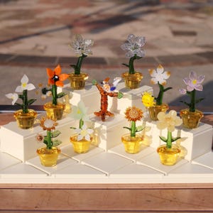 May include: A collection of 12 glass flower figurines in various colours, including white, orange, yellow, and pink, displayed on a white tiered stand. Each flower is potted in a clear glass pot.
